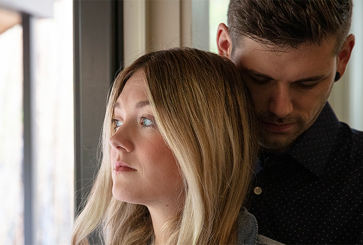 A couple stand by the window with a man hugging a woman from behind.