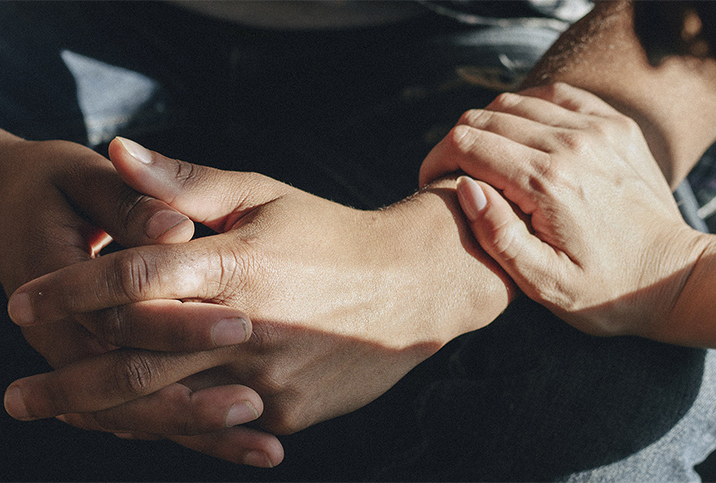 Two hands are folded together with another reaching out to hold his arm on the wrist.