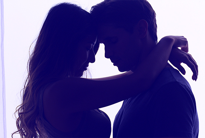 A couple stands in an embrace darkly lit with light behind them.