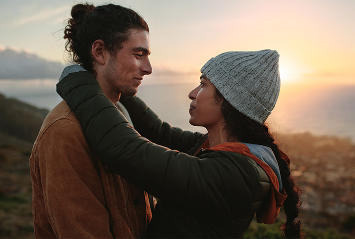 A man and a woman embrace cliffside in front of the sunset.