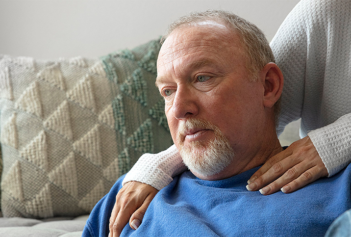 A man looks worried as he looks away with a woman resting her hands on his shoulders.