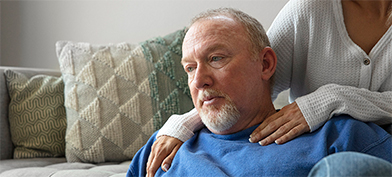 A man looks worried as he looks away with a woman resting her hands on his shoulders.
