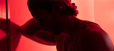 A man leans against a wall while holding his head with a red light shining behind him.