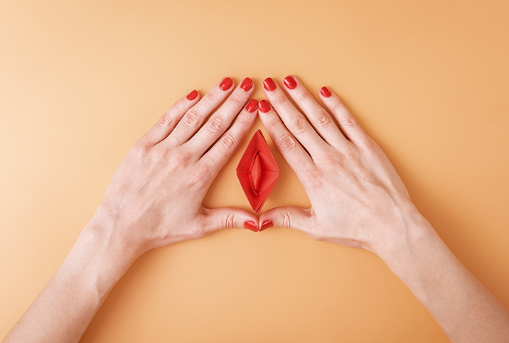 Two hands with red painted nails surround frame a vulva-shaped origami. 