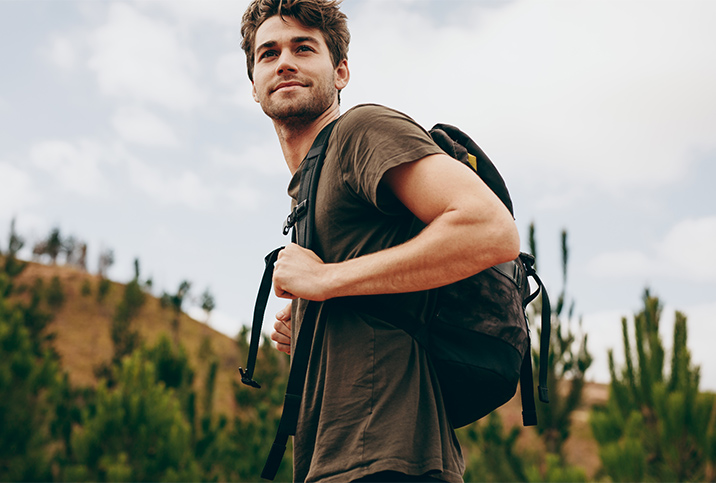 A young man wearing a backpack looks over his shoulder, smiling, with a mountain in the background.