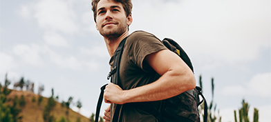 A young man wearing a backpack looks over his shoulder, smiling, with a mountain in the background.