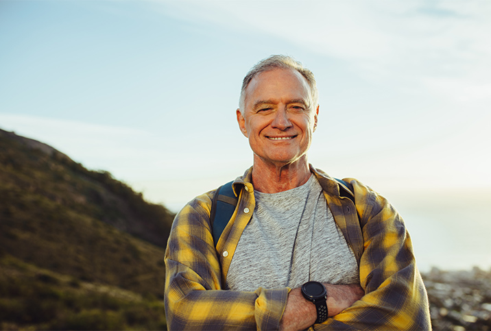 An older man is standing on a mountain and smiling at the camera with his arms folded.