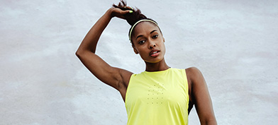 A woman in a yellow sleeveless shirt reaches to tug upwards on her ponytail.