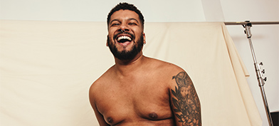 A shirtless man laughs in front of a white photography screen.