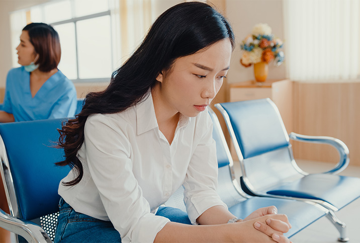 A woman sits in a medical waiting room looking down, concerned.