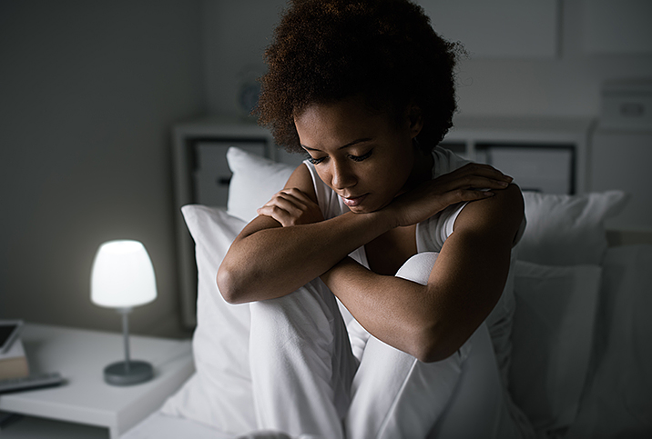 A grieving woman sits in bed with her legs up and arms folded over her knees.