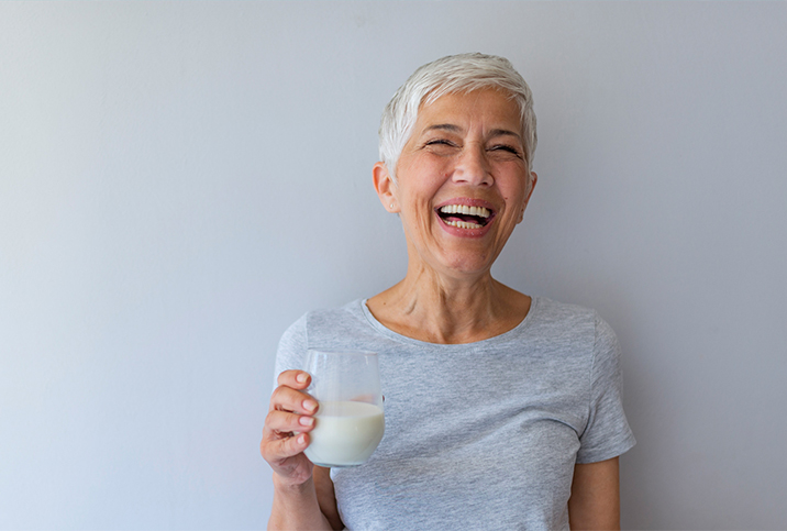 A woman with short white hair holds a glass of milk and laughs.