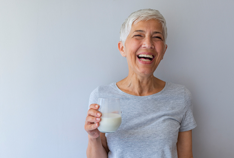 A woman with short white hair holds a glass of milk and laughs.