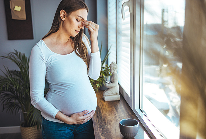 A pregnant woman with a migraine holds her belly and pinches her nose between her eyes.