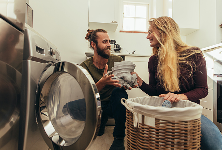 A woman hands her smiling partner laundry from the drying machine.