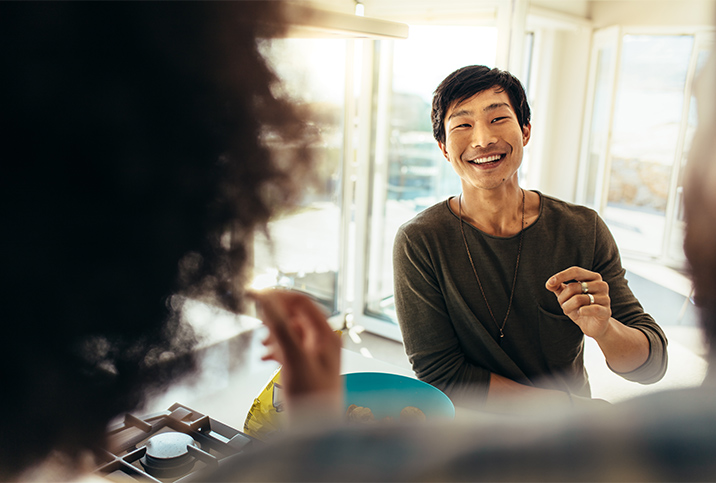 A man smiles at his friends in the kitchen as he brings food to his mouth. 