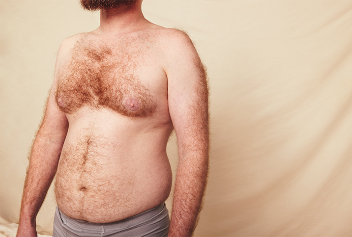 A shirtless man with a hairy chest and some belly fat stands in front of a beige background.