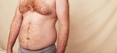 A shirtless man with a hairy chest and some belly fat stands in front of a beige background.