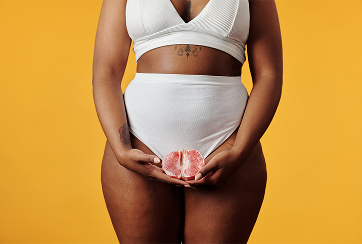 A woman in a white bra and high waisted underwear holds an open grapefruit in front of her crotch.