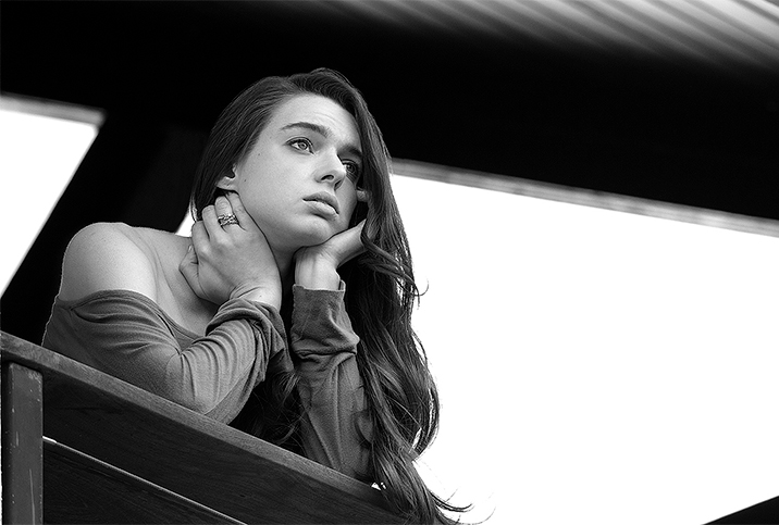 A woman rests her chin in her hands as she looks out from a balcony railing. 