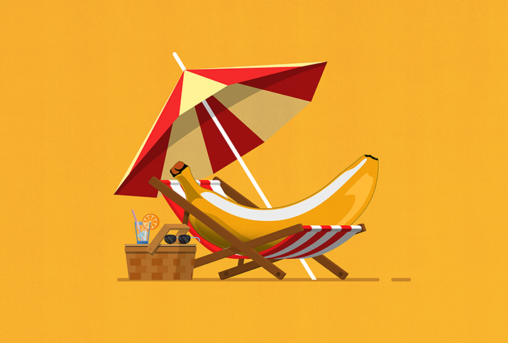 A banana lays in a beach chair underneath an umbrella and next to a picnic basket.