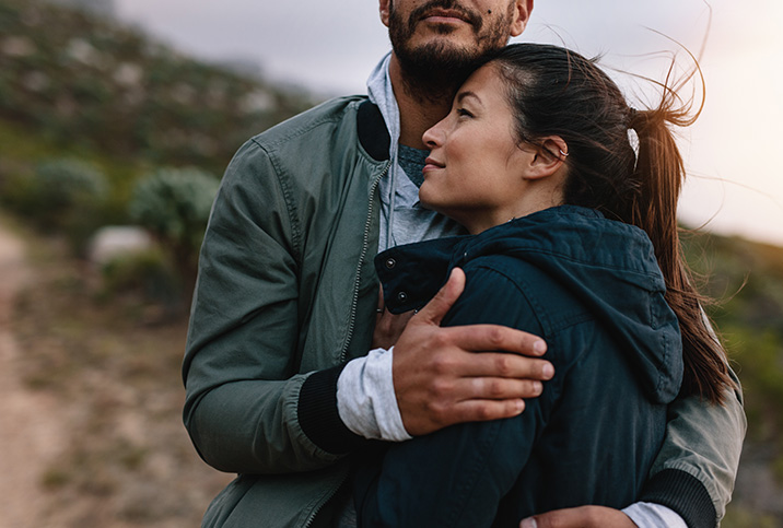 A couple stands outside, the woman leaning her head on a mans chest as he places his hand on her upper arm.