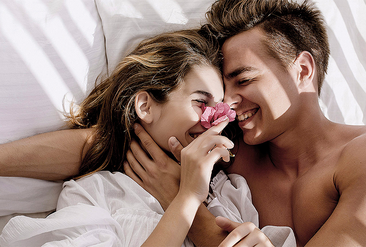 A man and woman cuddle in bed, their faces close together and a flower in the woman's hand.