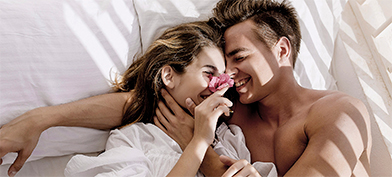 A man and woman cuddle in bed, their faces close together and a flower in the woman's hand.