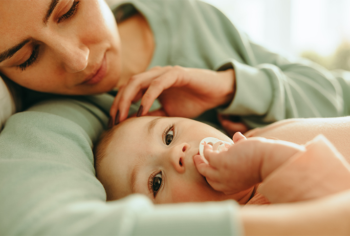 A mother touches her baby's head while it holds its pacifier and looks at the camera.