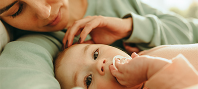 A mother touches her baby's head while it holds its pacifier and looks at the camera.