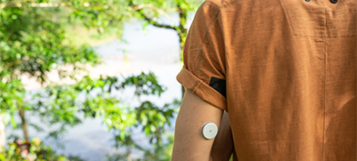 A man with diabetes stands near trees with his back to the camera with a glucose meter on his arm.
