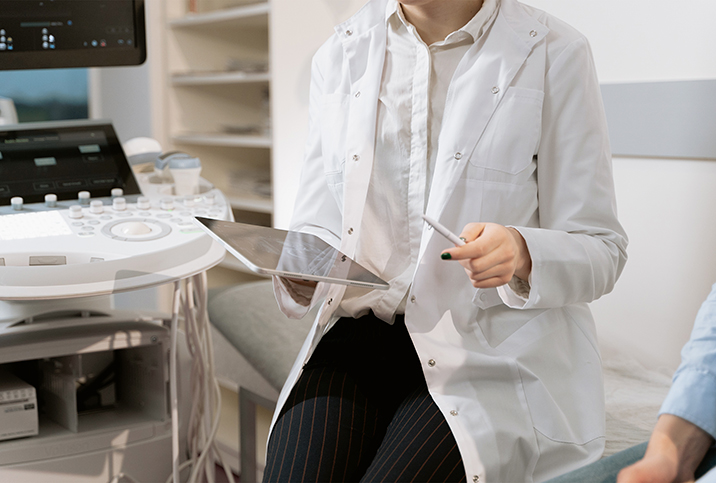 A doctor holding a tablet sits next to an ultrasound machine talking to a patient.