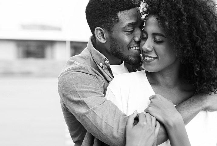 A smiling man wraps his arm around a smiling woman from behind.