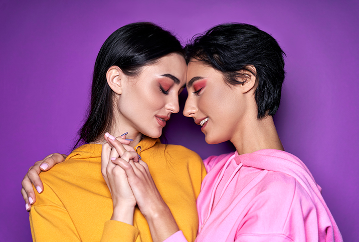 Two people with pink eye shadow press their foreheads together and hold hands in front of a purple background.