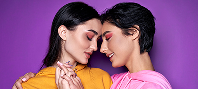 Two people with pink eye shadow press their foreheads together and hold hands in front of a purple background.