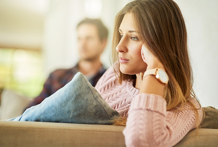 A woman stares blankly ahead with her head resting on her fist and her male partner sitting in the background.