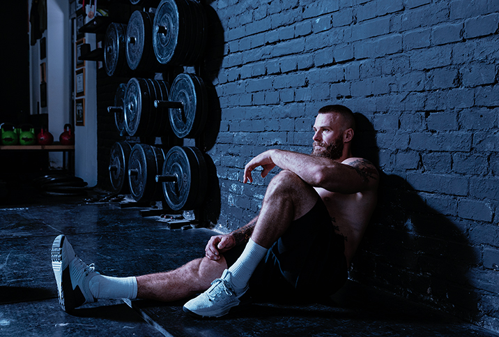 A bearded, shirtless man sits on the floor of a gym with his back against the wall. 