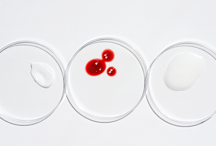 A petri dish with drops of period blood sits between two other petri dishes with skin care products.