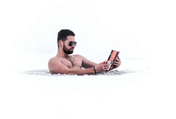 A man is doing cold water therapy outside in the opening of a snowy iced-over lake while reading a book.