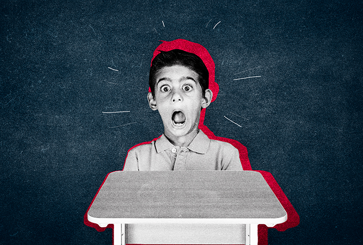 A little boy sits at a desk and makes a fearful expression with his eyes and mouth wide open.