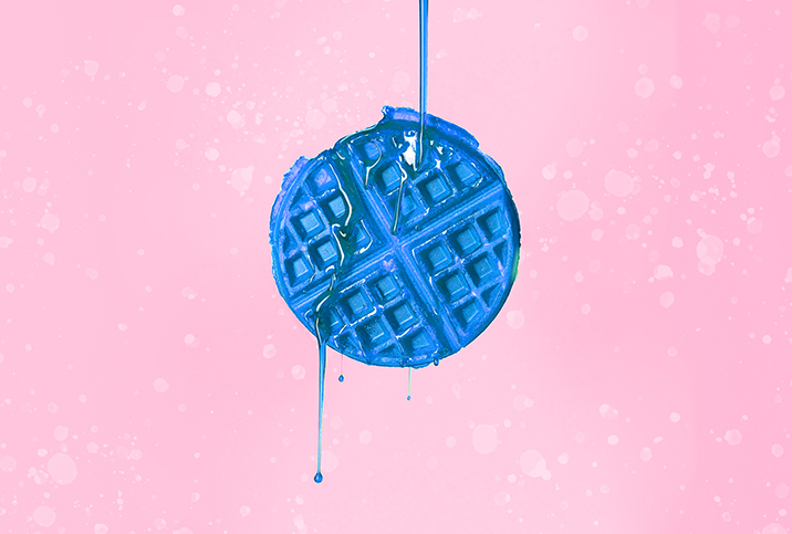 A blue waffle is against a pink background with blue syrup pouring down over it.