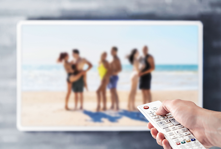 A hand holds a remote pointed at a television with a reality dating show on the screen.