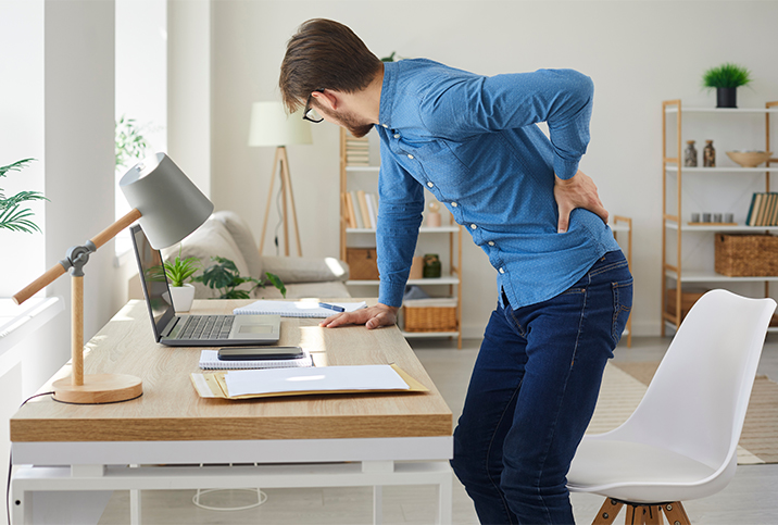 A man holds onto his lower back in pain while leaning on his computer desk.