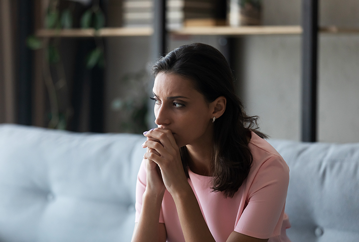 A concerned woman sits on a sofa with her hands held together against her chin.