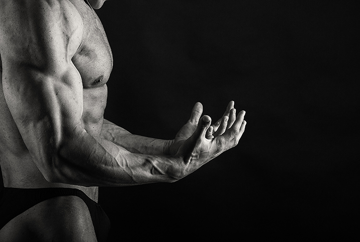 A muscled man flexes his arms out in front of him against a black background.