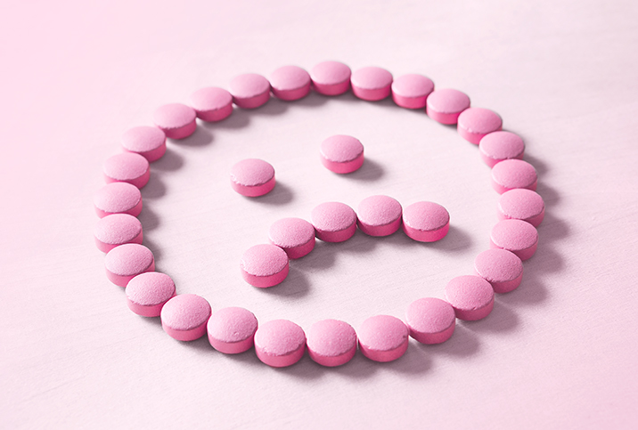 A frowning face made out of pink pills lays against a lighter pink surface.