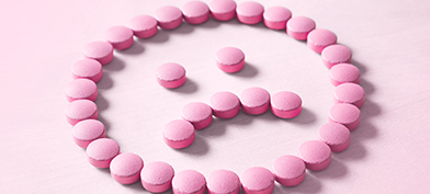 A frowning face made out of pink pills lays against a lighter pink surface.