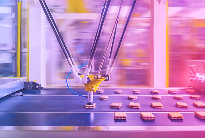 A manufacturing machine grabs a processed cookie sandwich from a conveyer belt.