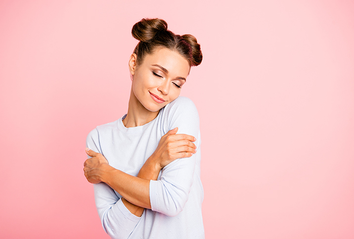 A person wearing their hair in buns closes their eyes and hugs themselves in front of a pink background.