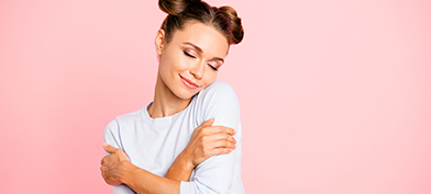 A person wearing their hair in buns closes their eyes and hugs themselves in front of a pink background.